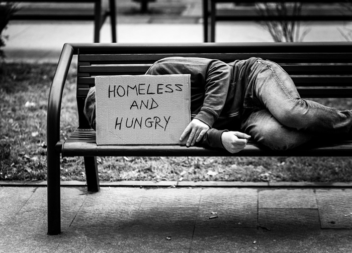 Person laying on bench with sign that reads HOMELESS AND HUNGRY